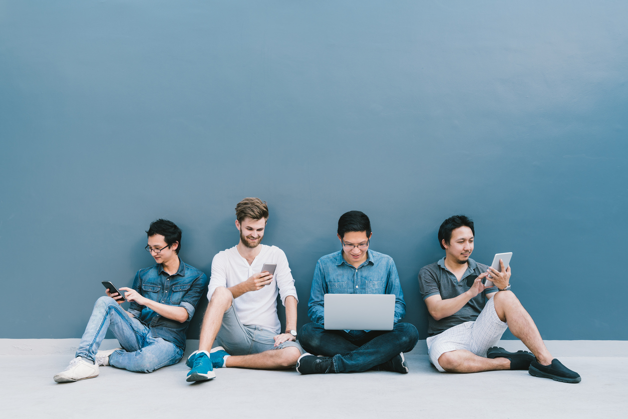 Multiethnic group of four men using smartphone, laptop computer, digital tablet together with copy space on blue wall. Lifestyle with infomation technology gadget, education, or social network concept