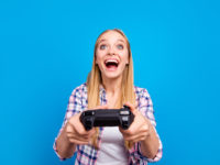 Close up of happy young blond girl holding joystick in hands and play in video games isoletad on blue background. Concept of victory and loses, competition and match